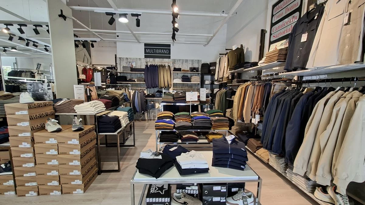 Interno del negozio Cortese Multibrand con giacche beige e blu appese, maglioni piegati su tavoli e scatole di scarpe