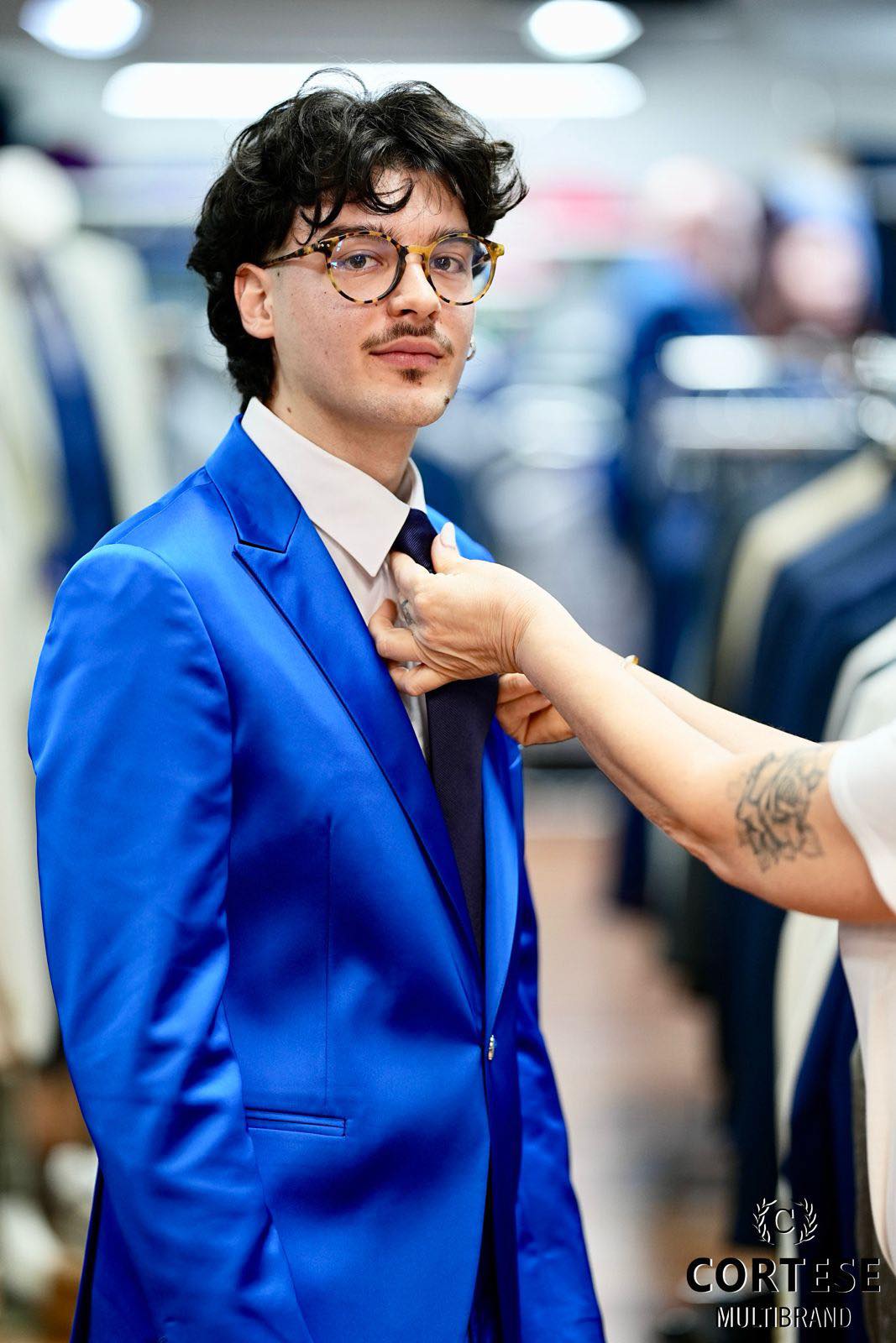 Ragazzo con occhiali tartarugati in giacca blu elettrico satinata, mani gli sistemano la cravatta da Cortese Multibrand